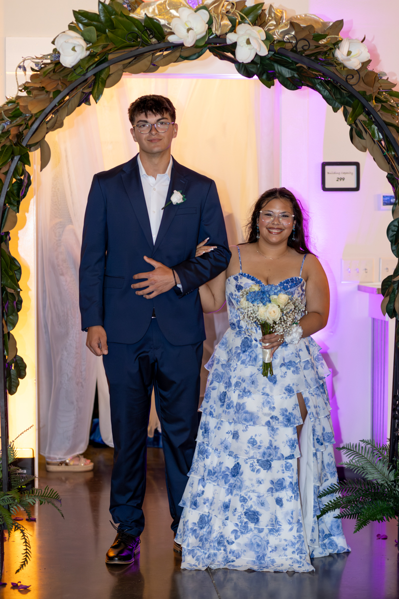 Couple posing under a flower-covered arch at a formal event; man in a navy suit with a white boutonniere, woman in a blue floral gown holding a bouquet.