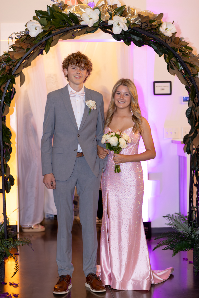 Couple posing under a flower arch at a formal event; man in a light gray suit and bow tie, woman in a pink glitter dress holding a bouquet.