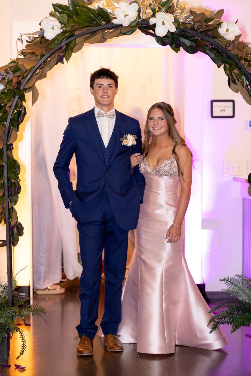 Couple in formal attire posing arm-in-arm under a flower arch at a decorated event venue.