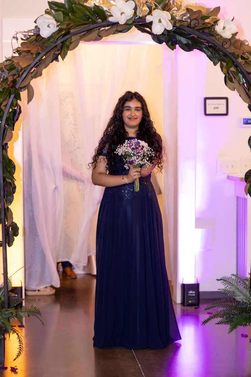 Young woman in a navy blue evening gown holding a bouquet, standing under a floral arch at a formal event.