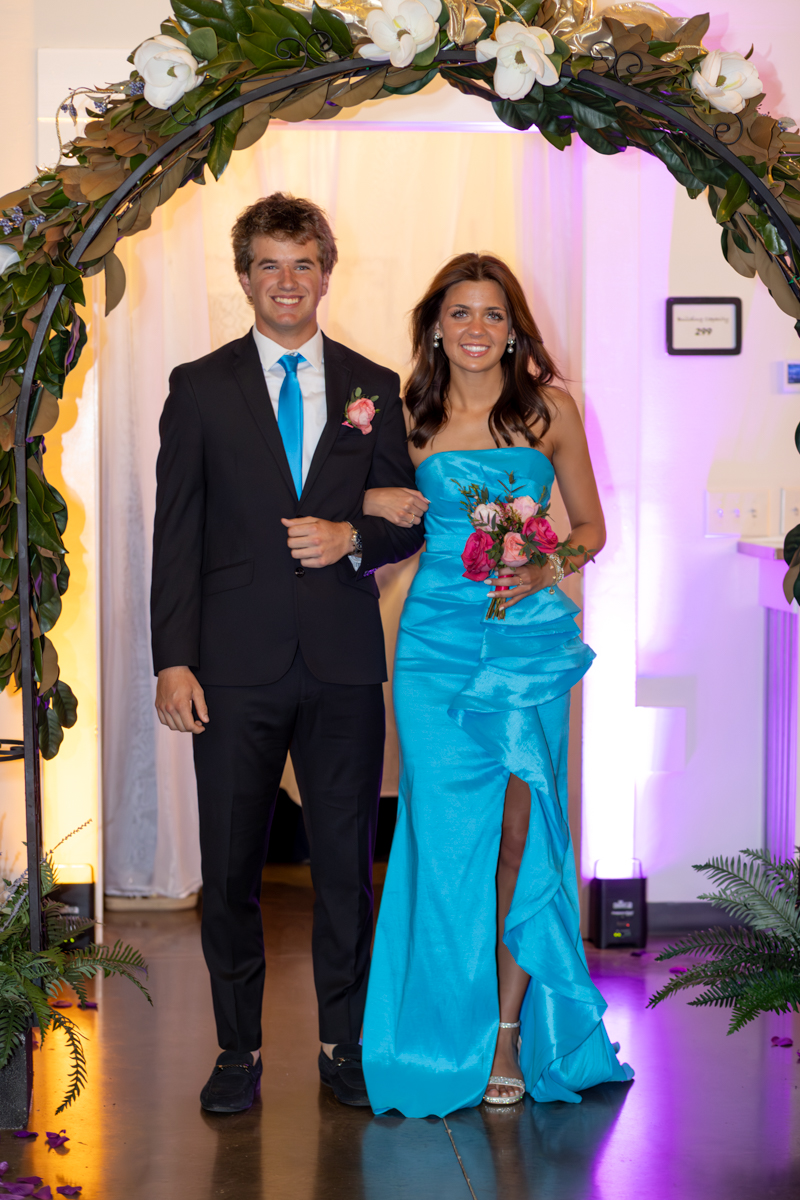 Smiling couple walking under a floral arch at a formal event; man in a black suit with blue tie, woman in turquoise gown holding pink bouquet.