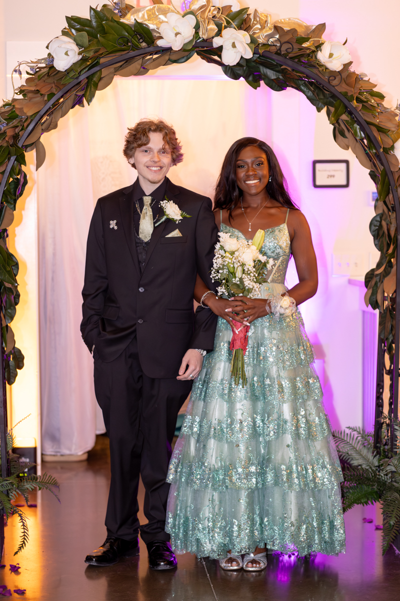 Couple posing under a large floral arch at a formal event: the man in a black suit and tie, the woman in a sparkling turquoise gown holding a bouquet, pink and purple lighting in the background.