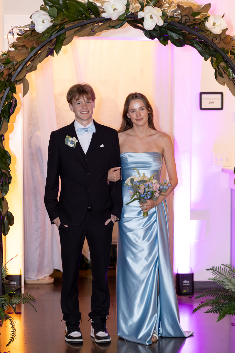 Couple in formal attire posing under a floral arch: man in black suit with a light-blue bow tie and boutonniere, woman in a blue satin strapless gown holding a bouquet, pink/purple uplighting nearby.