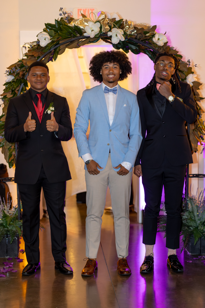 Three young men in formal wear stand under a flower arch at an event; left and right wear dark suits, center wears a light blue blazer and bow tie, all smiling.