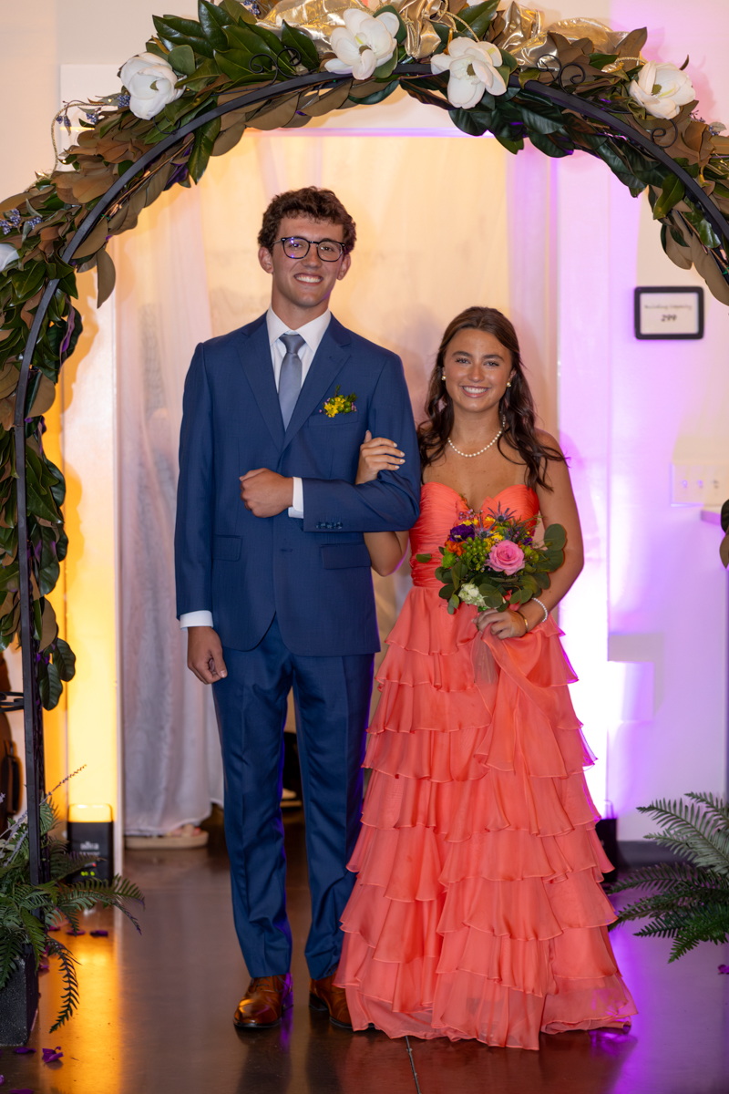 Smiling couple in formal attire walking under a flower arch at a formal event.