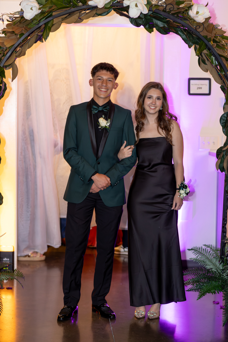 Two teens in formal attire pose arm-in-arm beneath a flower-covered arch at a formal event.