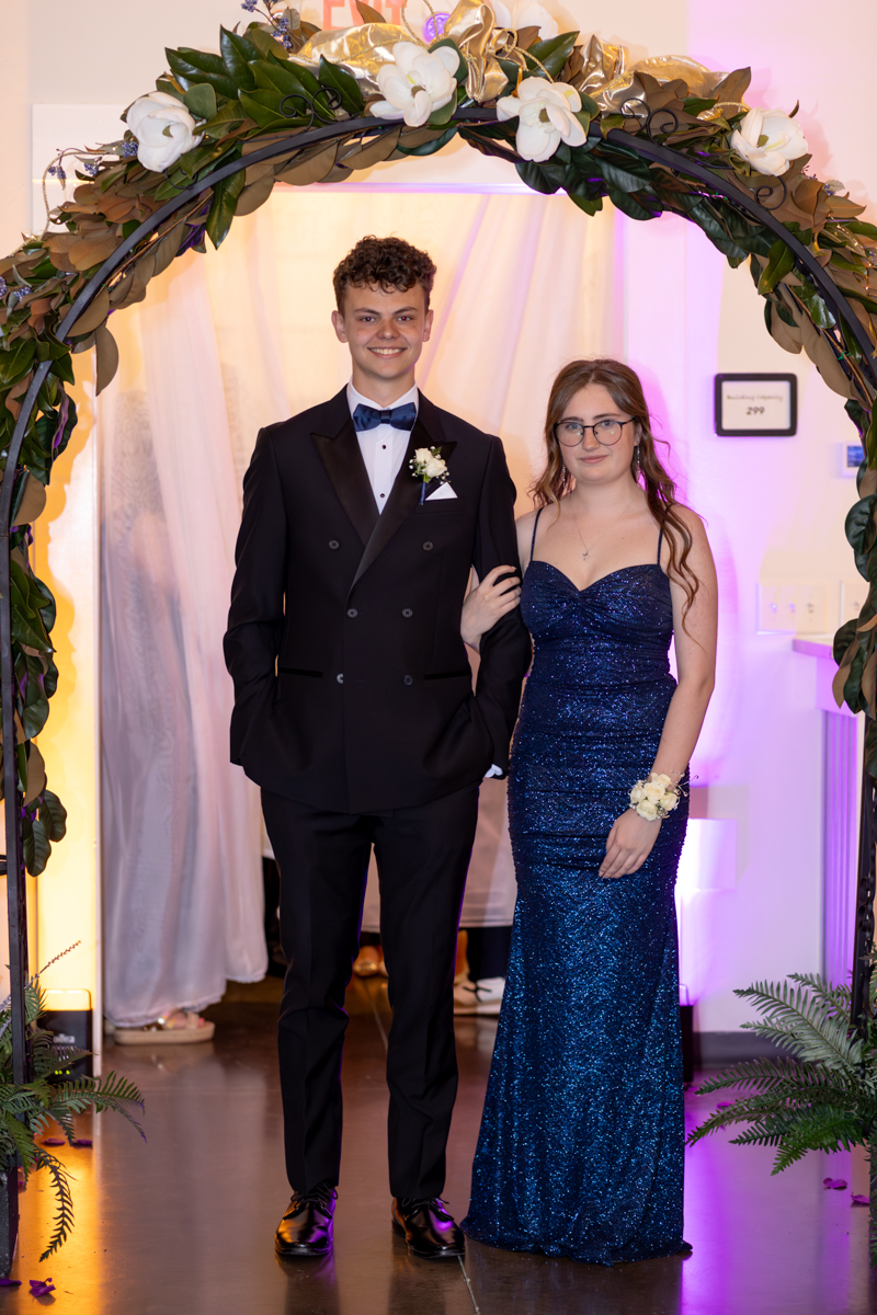 Two teens in formal wear pose arm-in-arm under a flower arch at a formal event; the boy wears a black tuxedo and bow tie, the girl a blue sequin gown with a corsage.