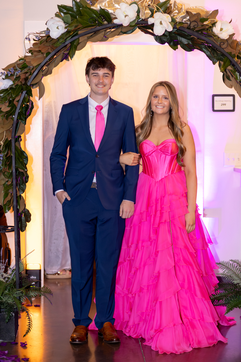 Couple posing under a flower arch at a formal event; man in a navy suit with a pink tie and the woman in a bright pink tiered gown.