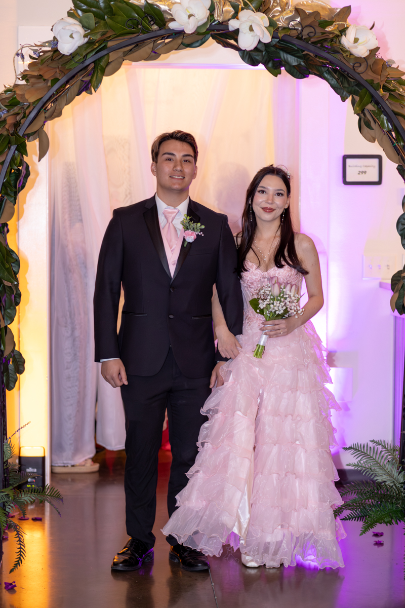 Couple posing under a floral arch at a formal event; man in a dark suit with pink tie, woman in a pink layered gown holding a small bouquet.