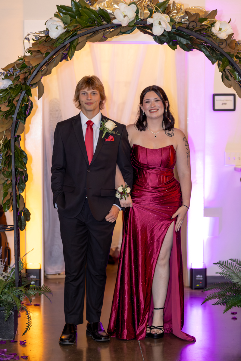 Young couple in formal attire posing arm-in-arm under a flower-adorned arch, the man in a black suit with red tie and boutonniere, the woman in a magenta sequined gown with a thigh-high slit.