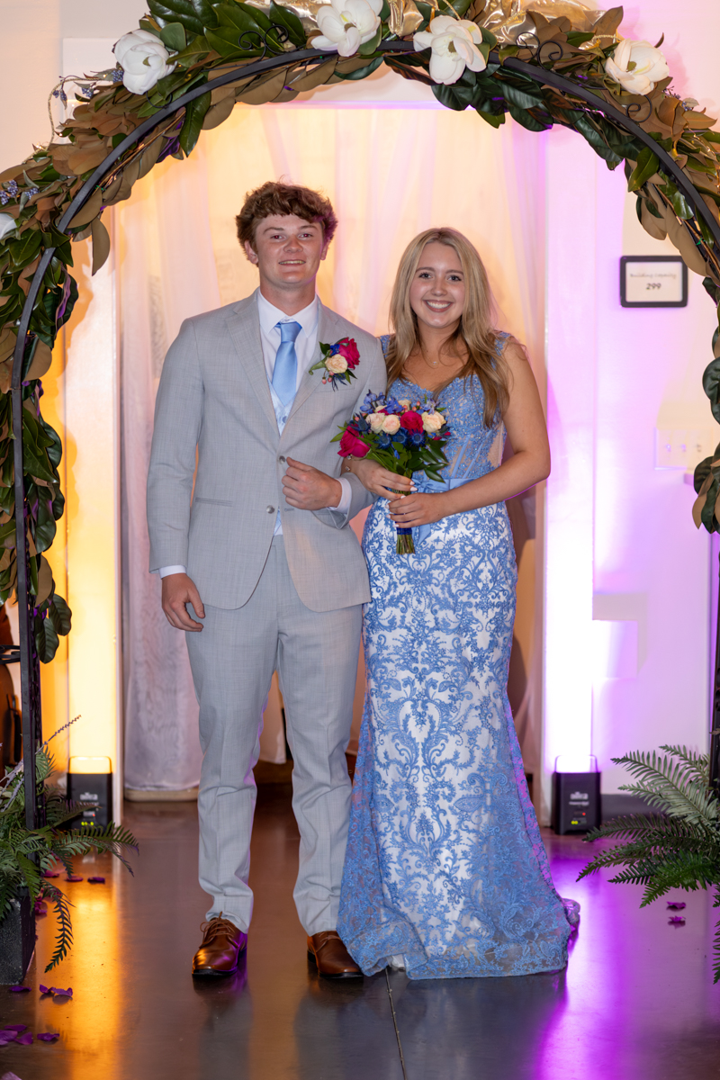 A young couple walking under a flower arch at a formal event; the man in a light gray suit with blue tie and boutonniere, the woman in a blue lace gown holding a bouquet.