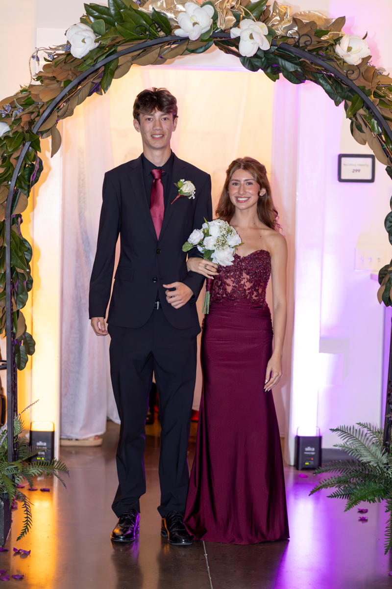 Couple posing under a floral arch: tall man in a black suit with red tie and boutonniere, woman in a burgundy gown holding a bouquet of white flowers.