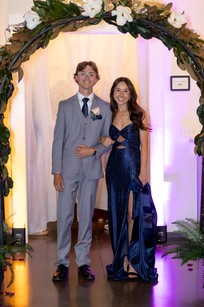 A young couple posing under a floral arch at a formal event; man in a light gray suit with boutonniere, woman in a navy sequined gown.
