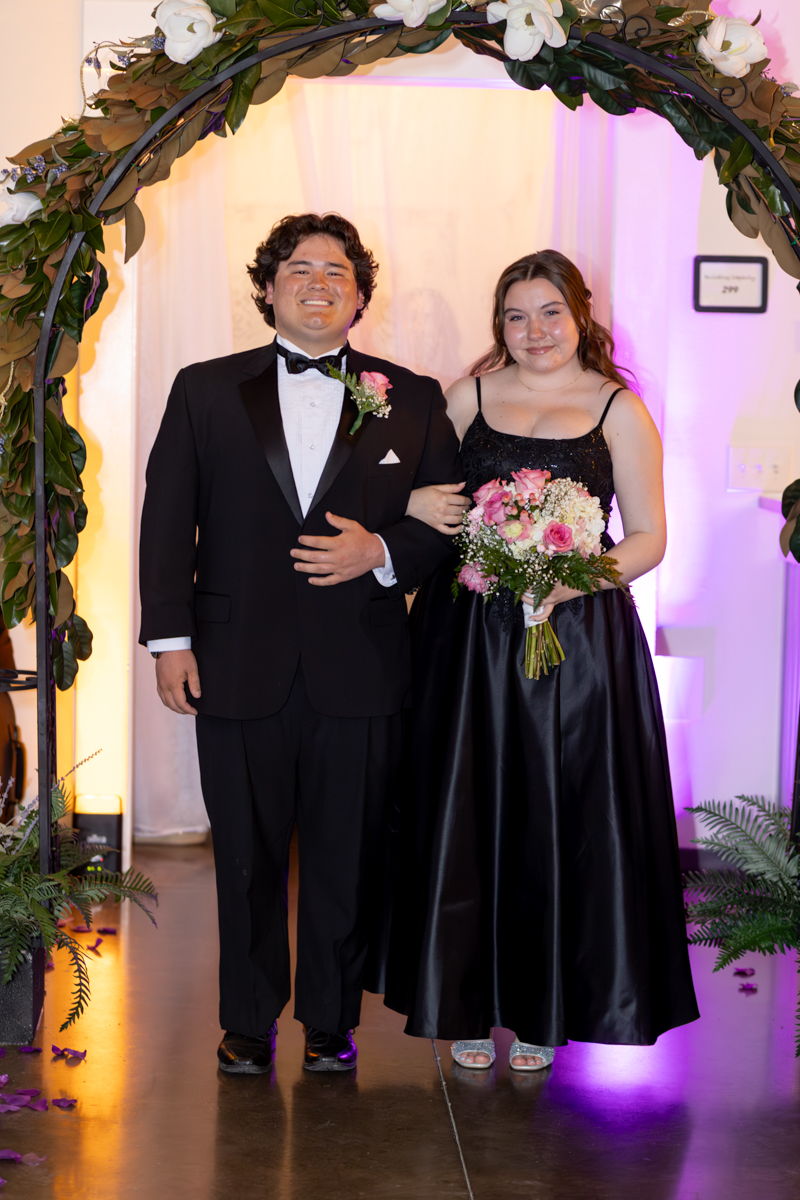 Young couple in formal attire posing under a flower arch; man in a tuxedo with a pink boutonniere, woman in a black gown holding a bouquet.