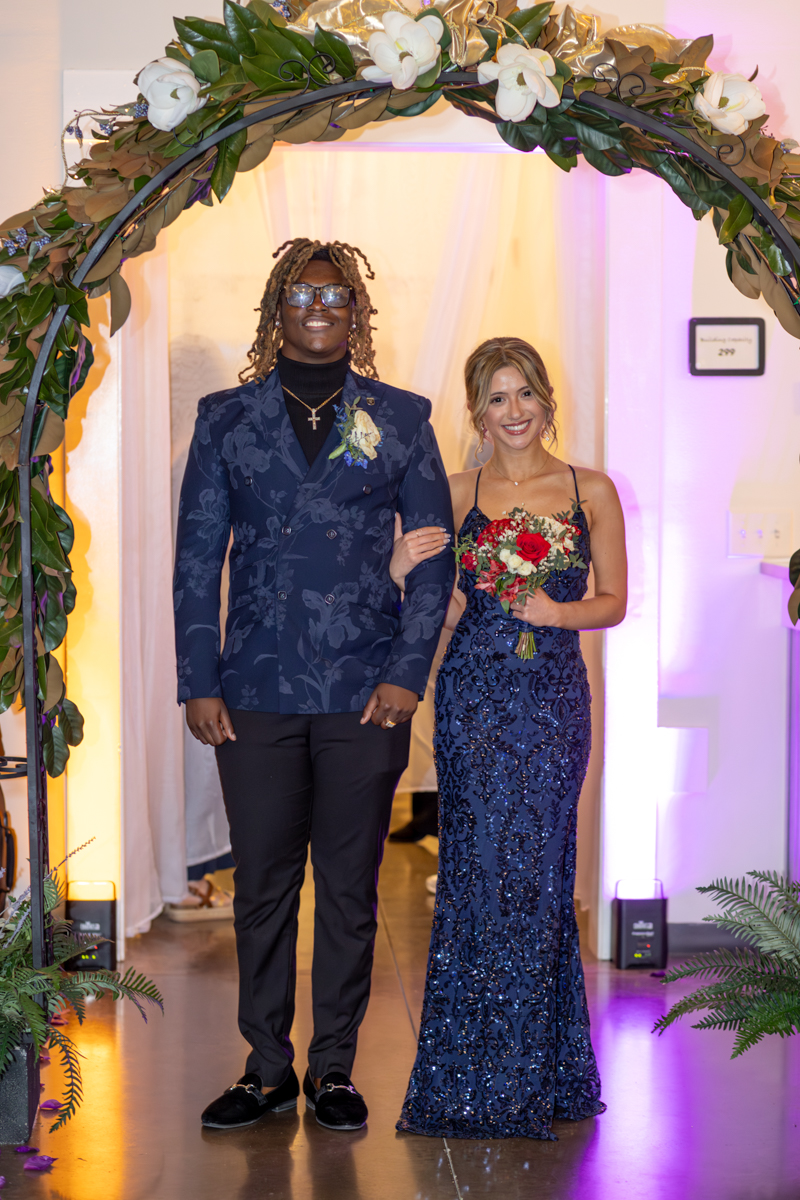 Couple walks arm in arm under a flower-adorned arch at a formal event; man wears a navy floral blazer, woman in a beaded navy gown holding a bouquet.