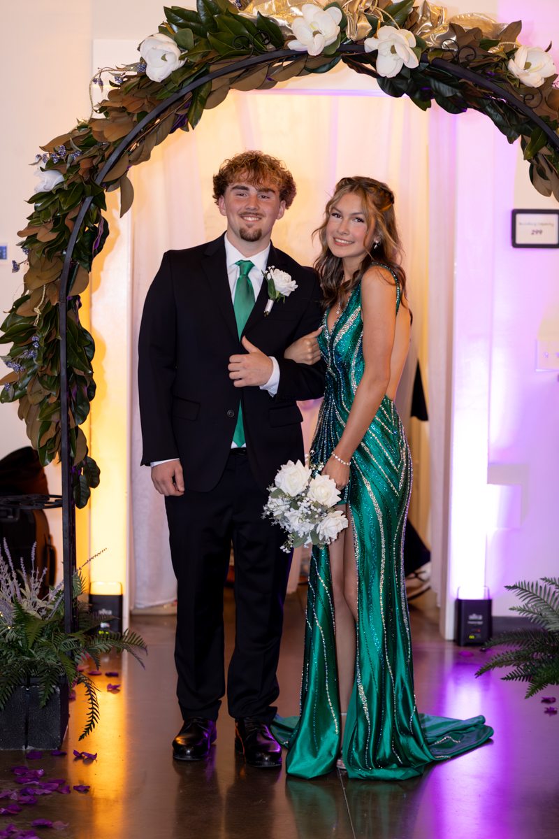 Couple in formal wear posing under a flower arch; man in a black suit with green tie, woman in an emerald beaded gown holding a white bouquet.