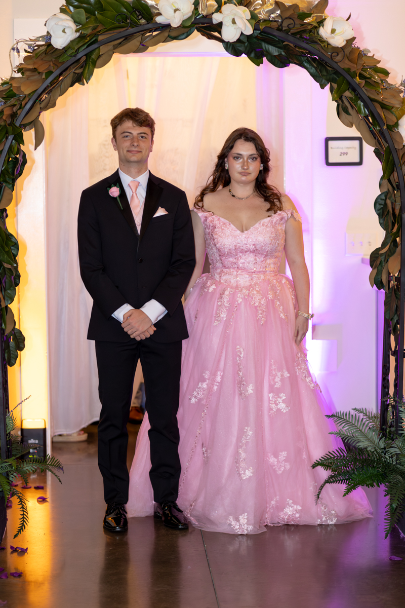 Young man in a black suit with pink tie and boutonniere beside a woman in a pink lace evening gown under a flowered arch at a formal event.