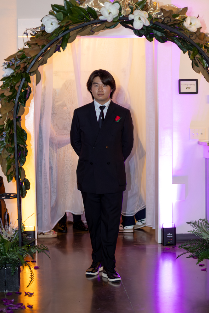 Man in a dark suit with a red boutonniere standing under a floral arch at an indoor event, with purple lighting and white curtains behind him.