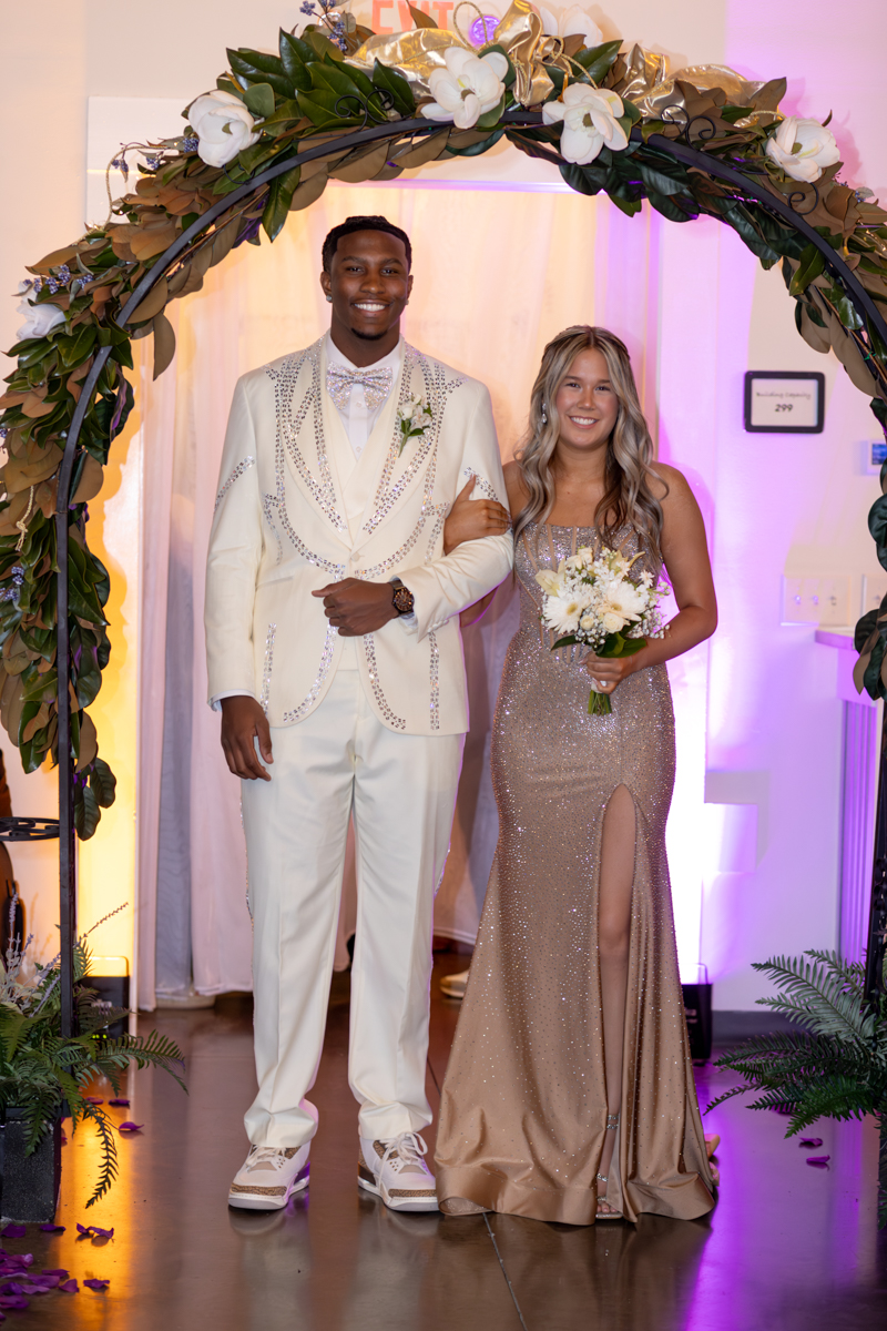 Couple posing under a flower arch at a formal event; man in a white embellished suit and bow tie, woman in a sparkling champagne gown holding a bouquet in arm-in-arm stance.