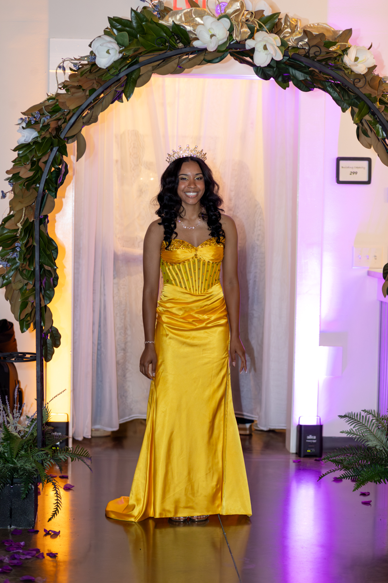 Woman in a bright yellow satin gown with a tiara standing under a floral arch at a formal event.
