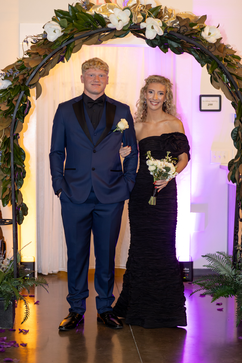 Couple posing under a floral arch at a formal event; man in navy tuxedo with boutonniere, woman in a black off‑the‑shoulder gown holding a bouquet.