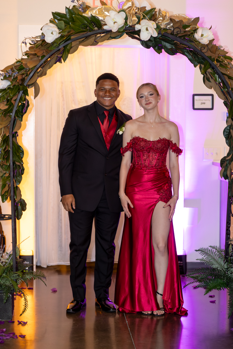 Couple posing under a floral arch at a formal event; man in a black suit with red tie, woman in a red off-shoulder gown with a high slit.