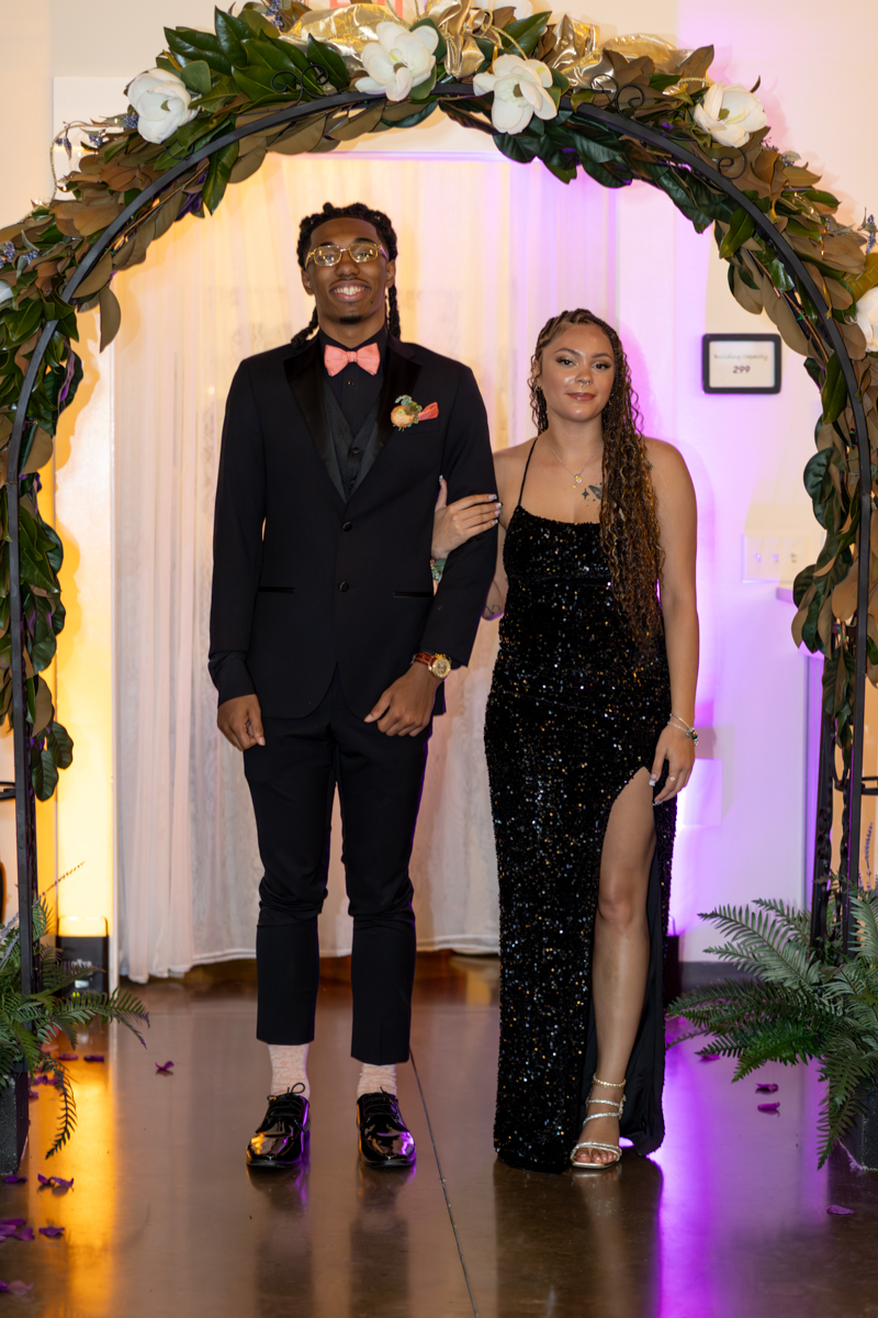 Couple in formal wear posing under a flower arch at a decorated event venue.