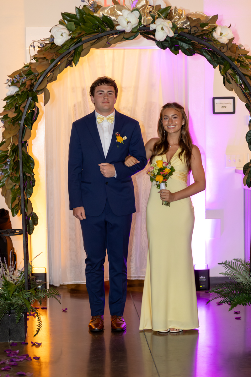 Couple walking arm-in-arm under a flower arch at a formal event; man in navy suit with yellow bow tie and boutonniere, woman in pale yellow dress holding a bouquet.