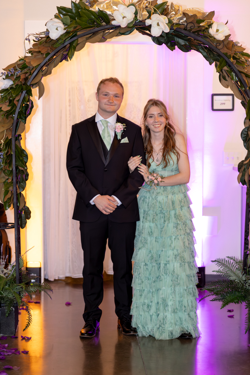 Young couple in formal attire posing under a flower-covered arch at a ceremony or prom, the man in a black suit with mint-green tie and boutonniere, the woman in a mint-green tiered dress holding his arm.