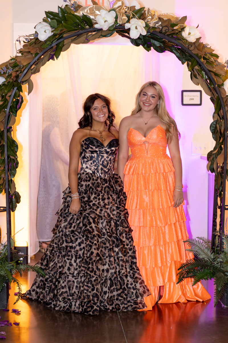 Two women in formal gowns pose under a floral arch at a formal event.