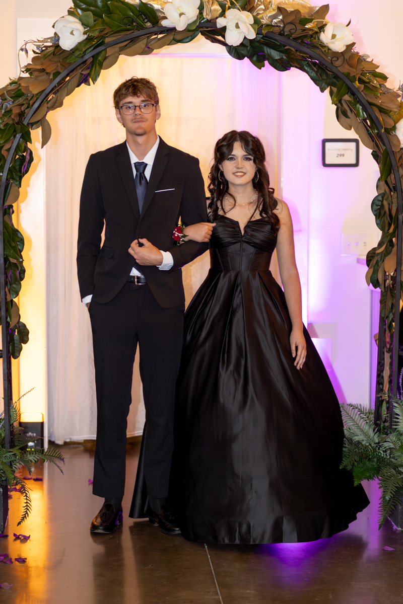 Two young adults in formal wear walk through a floral arch, the man in a suit and the woman in a black ball gown.
