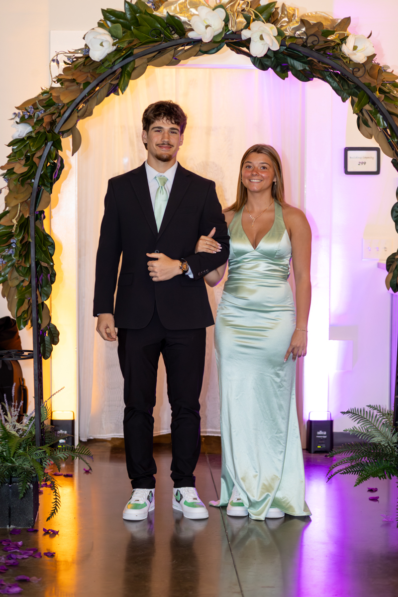 Couple under a floral arch at a formal event: man in a black suit with a mint tie and woman in a mint-green satin gown walking arm-in-arm.",