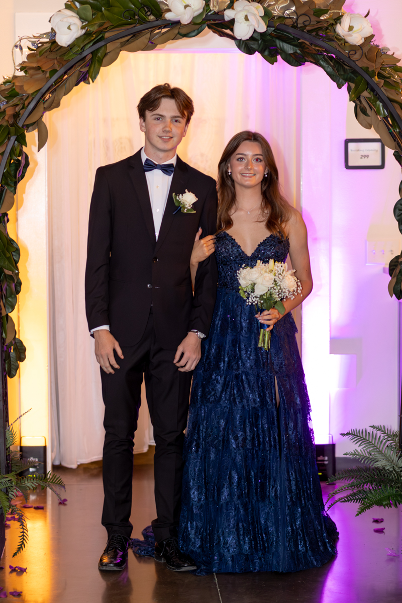 A couple posing under a floral arch at a formal event; man in a black suit with a bow tie, woman in a navy blue gown holding a white bouquet.