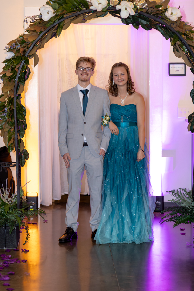Two teenagers in formal attire walk arm in arm under a flower-covered arch at a decorated event venue.