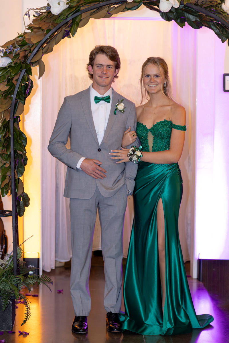 Young couple posing under a floral arch at a formal event; man in a light gray suit with a green bow tie, woman in an emerald green gown with a thigh-high slit.