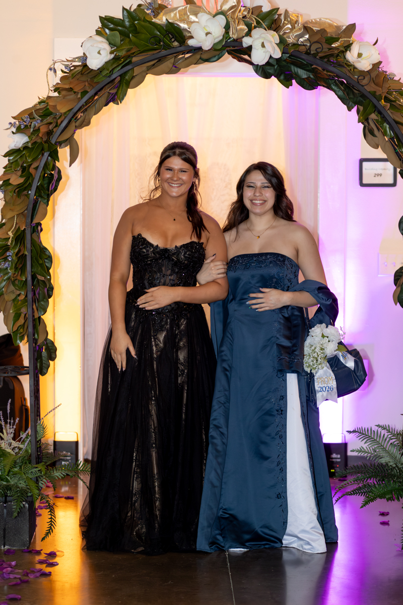 Two young women in formal gowns smile and pose under a floral arch at a prom or formal event, arm in arm in front of a lit backdrop.