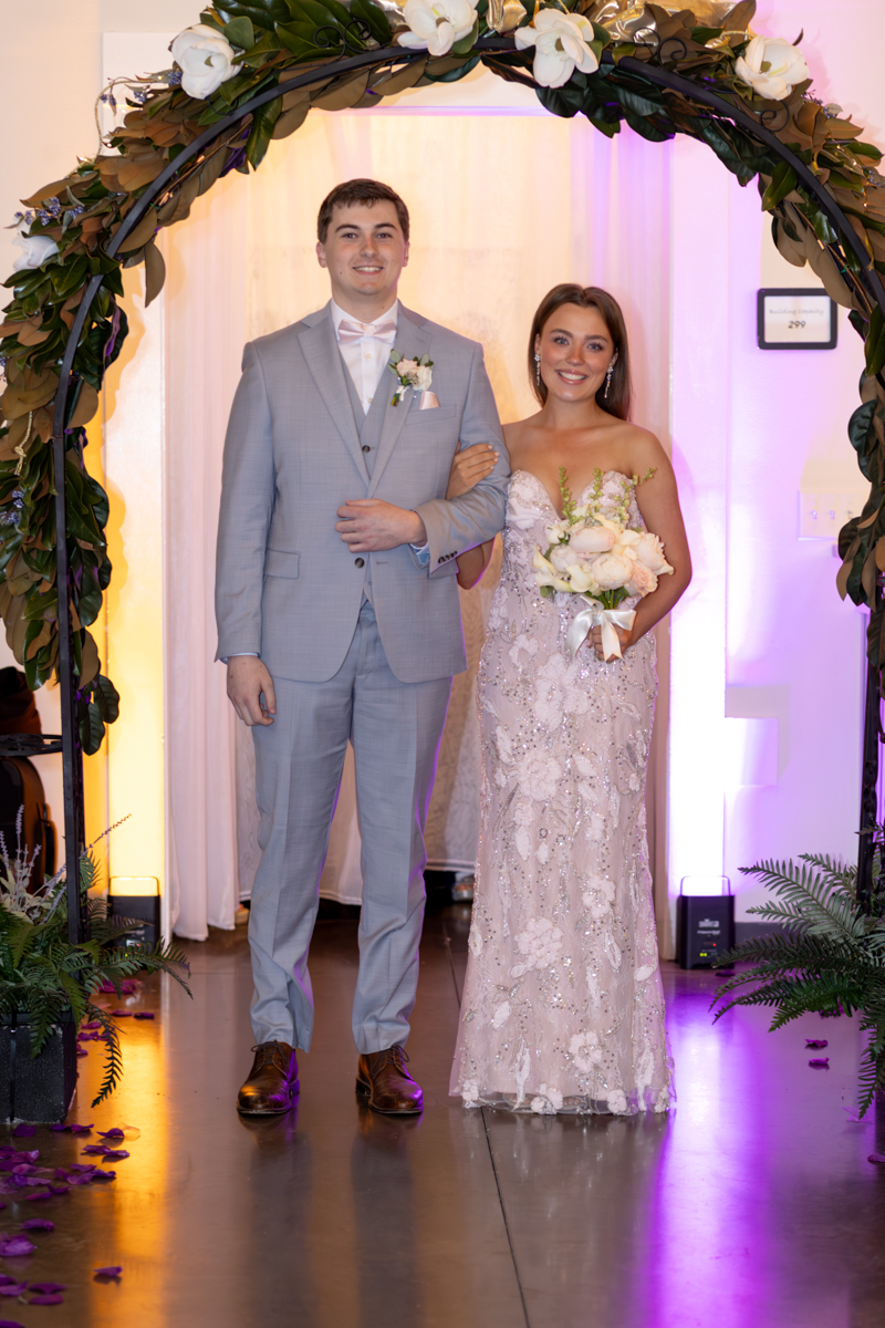 Bride and groom walk arm-in-arm under a floral arch, the bride in a beaded gown holding a white bouquet.