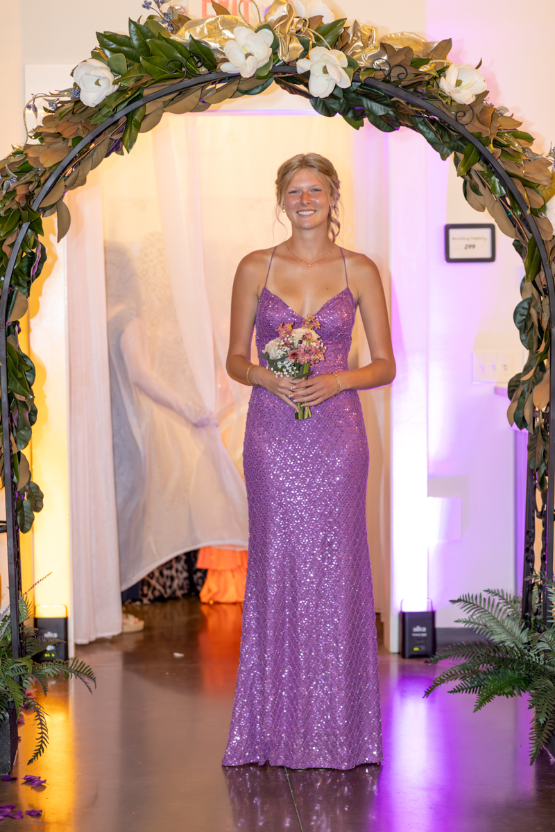 Smiling woman in a lavender sequin gown holding a small bouquet under a flower-adorned arch at a formal event=wreath arch with white blooms and greenery