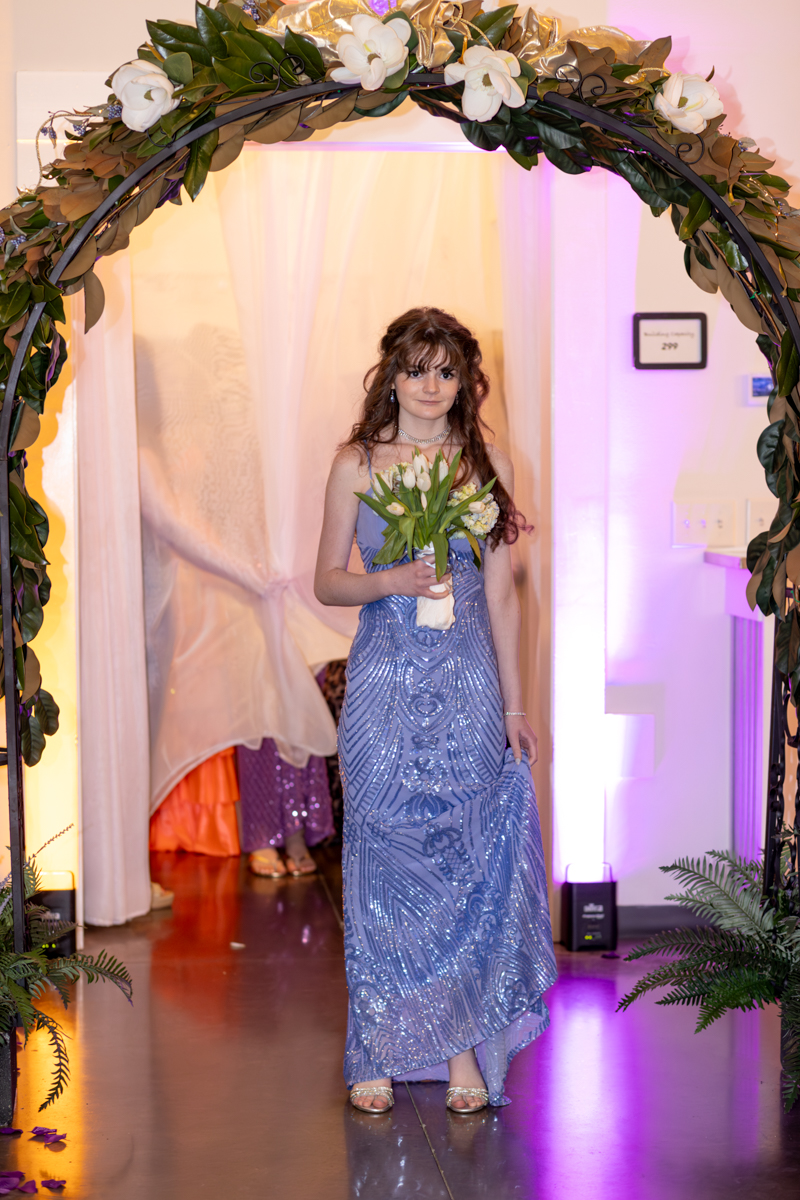 Young woman in a shimmering lavender gown walks through a flower arch, holding a bouquet of white tulips.