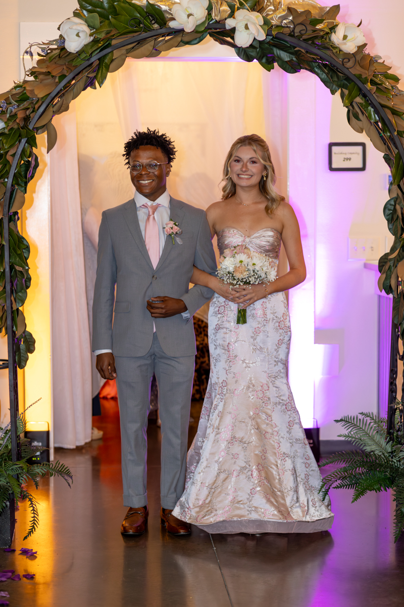 Bride and groom walk arm in arm under a flower arch at their wedding; he wears a gray suit with a pink tie, she wears a pastel lace gown holding a bouquet.