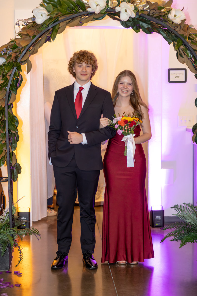Young couple in formal wear walk arm-in-arm under a floral arch, the woman in a burgundy gown holding a bouquet with a white ribbon.