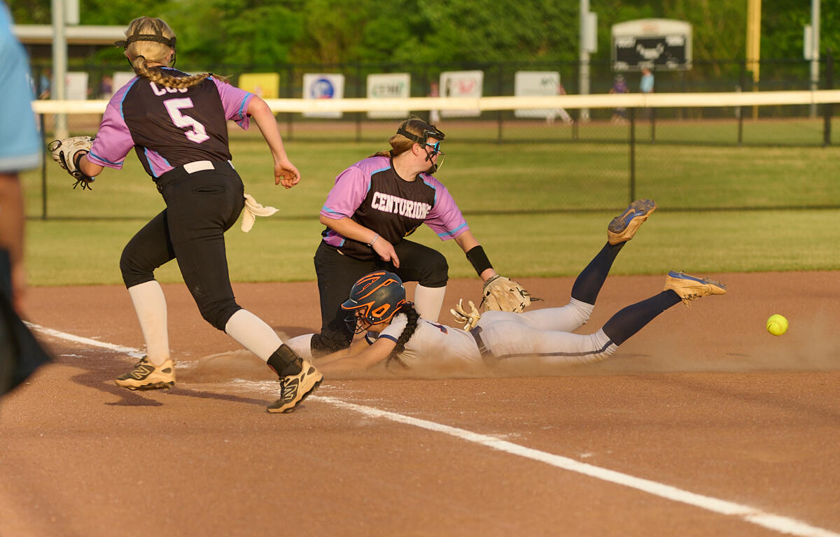 Softball infield action: a baserunner slides headfirst into the dirt as a fielder extends to reach the ball behind her, with teammates nearby.