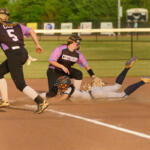 Softball infield action: a baserunner slides headfirst into the dirt as a fielder extends to reach the ball behind her, with teammates nearby.
