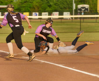 Softball infield action: a baserunner slides headfirst into the dirt as a fielder extends to reach the ball behind her, with teammates nearby.