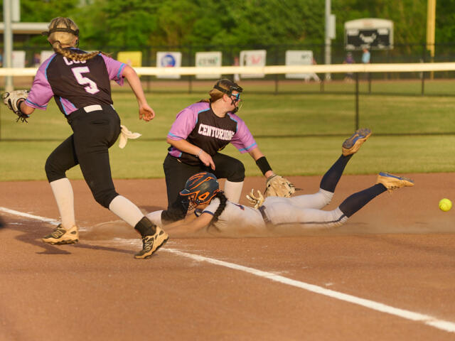 Softball infield action: a baserunner slides headfirst into the dirt as a fielder extends to reach the ball behind her, with teammates nearby.