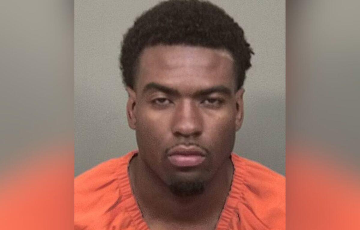 Mugshot of a young man wearing an orange jail jumpsuit, facing the camera with a neutral expression against a gray background from chest up.