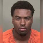 Mugshot of a young man wearing an orange jail jumpsuit, facing the camera with a neutral expression against a gray background from chest up.