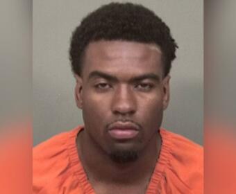 Mugshot of a young man wearing an orange jail jumpsuit, facing the camera with a neutral expression against a gray background from chest up.