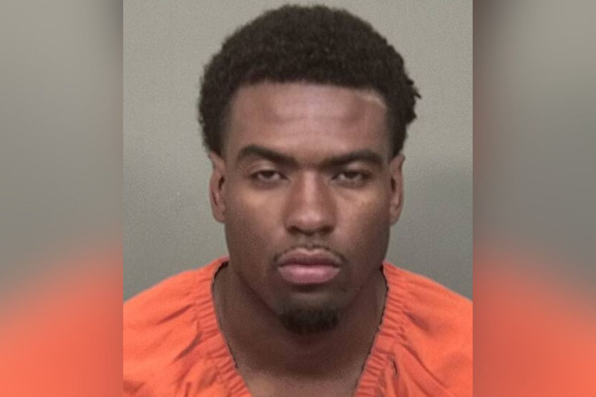 Mugshot of a young man wearing an orange jail jumpsuit, facing the camera with a neutral expression against a gray background from chest up.
