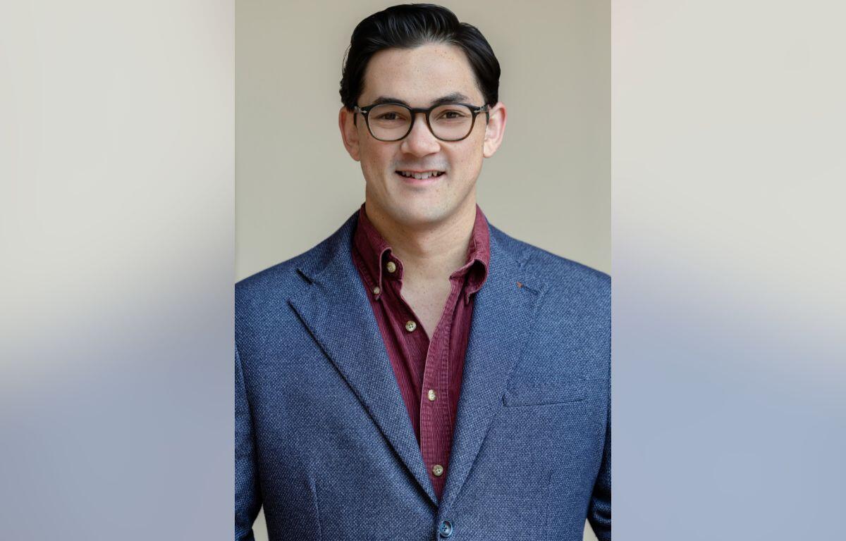 Smiling man wearing glasses and a blue blazer over a maroon shirt against a neutral background.